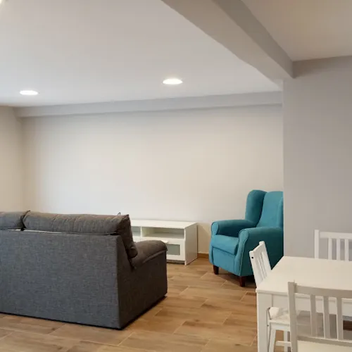 Sala de estar de una casa donde se está un sofá gris y otro azul turquesa. Además hay una mesa blanca de madera con sus sillas a juego y un mueble de salón para sujetar la tele.