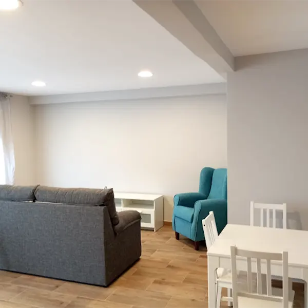 Sala de estar de una casa donde se está un sofá gris y otro azul turquesa. Además hay una mesa blanca de madera con sus sillas a juego y un mueble de salón para sujetar la tele.